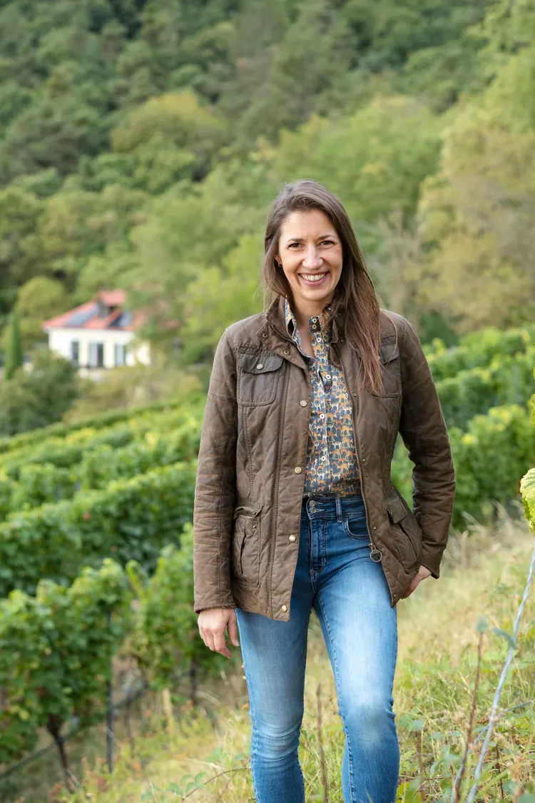 Winzerin Victoria Lergenmüller steht lachend zwischen den Rebzeilen, im Hintergrund das Gutshaus am Waldrand