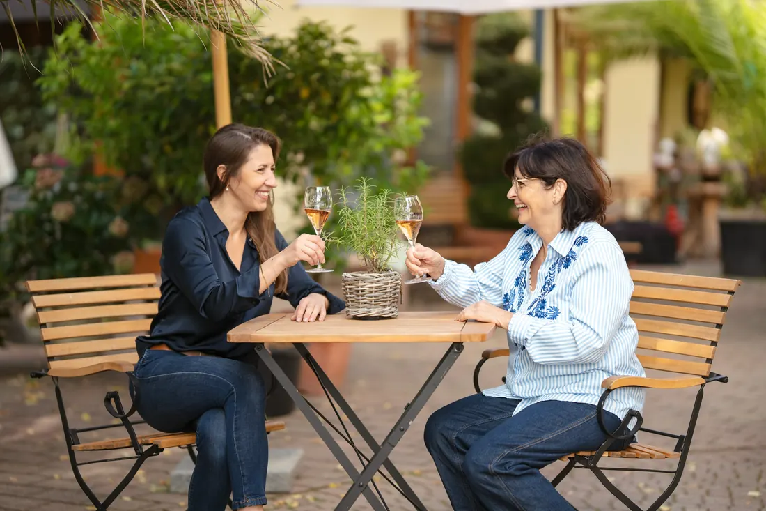Zwei Frauen stoßen auf der Terrasse an einem kleinen Holztisch mit einem Glas Wein an